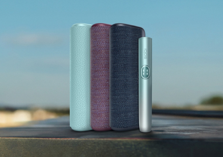 Três equipamentos IQOS ILUMA i PRIME na cor azul claro, vermelho escuro, azul escuro e dispositivo azul claro em ambiente de exterior desfocado
