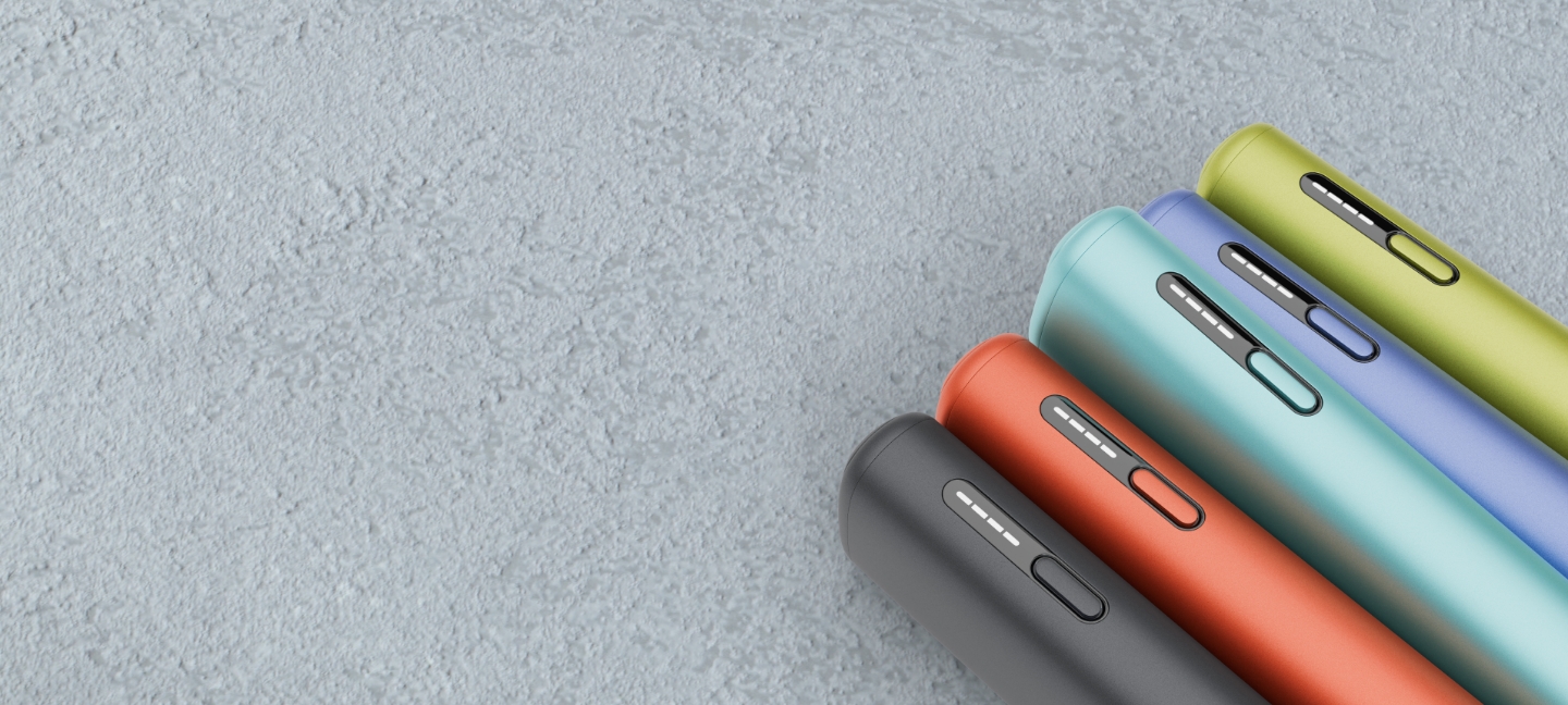 Cinco equipamentos IQOS ILUMA i ONE na cor azul escuro, terracota, azul claro, violeta e verde, com detalhe das luzes de presença, no  canto inferior direito da imagem e orientados para o canto superior esquerdo, em ambiente de cimento