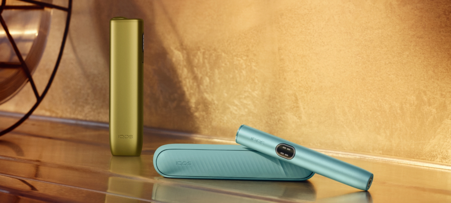 Equipamento IQOS ILUMA i ONE verde na vertical e equipamento IQOS ILUMA i ONE azul claro pousado na horizontal, com dispositivo inclinado sobre o carregador de bolso,em ambiente luminoso com decoração.