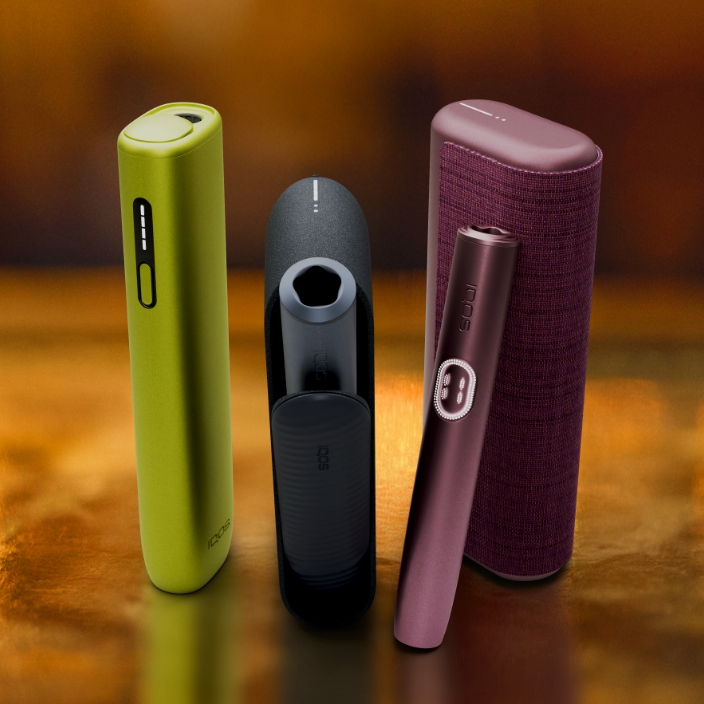 Equipamento IQOS ILUMA i ONE verde orientado para a esquerda, equipamento IQOS ILUMA i azul escuro ao centro e equipamento IQOS ILUMA i PRIME vermelho escuro com dispositivo encostado ao carregador de bolso orientado para a direita, em fundo luminoso