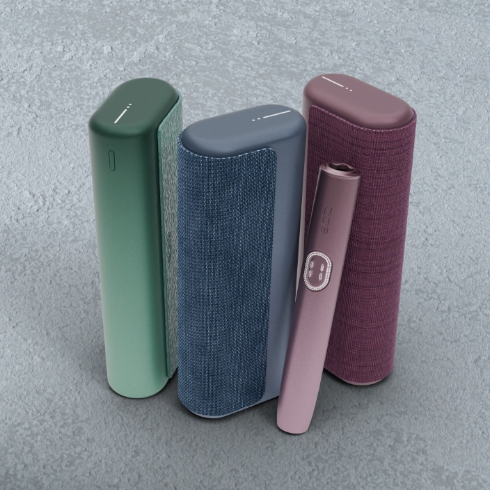 Equipamento IQOS ILUMA i PRIME verde escuro orientado para a esquerda, equipamento IQOS ILUMA i PRIME azul escuro ao centro e equipamento IQOS ILUMA i PRIME vermelho escuro com dispositivo encostado ao carregador de bolso e orientado para a direita, em fundo cimento