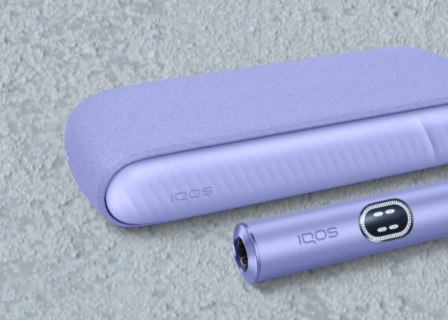 Detalhe de equipamento IQOS ILUMA i violeta, orientado para a esquerda com destaque do carregador de bolso e luzes de presença do dispositivo, em ambiente de cimento.