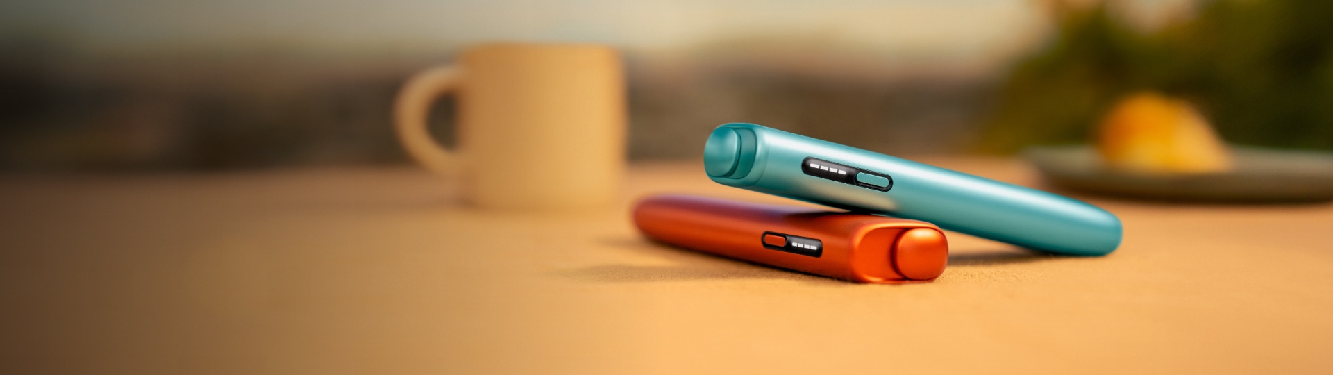 Dois dispositivos, IQOS ILUMA i ONE Azul Claro sobreposto a dispositivo IQOS ILUMA i ONE Vermelho, sobre mesa