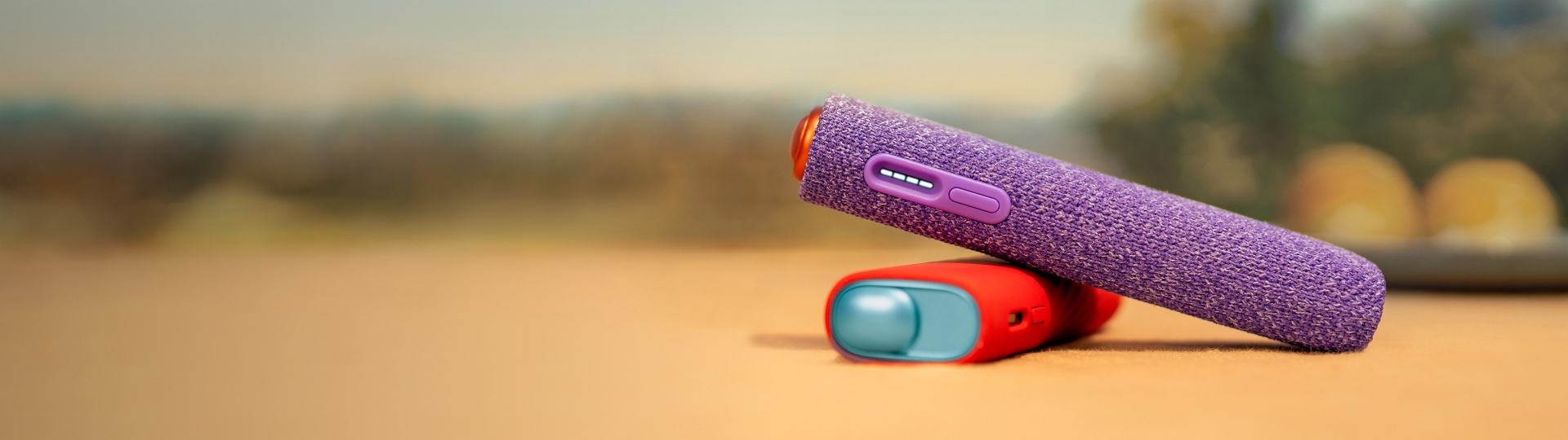 Dispositivo IQOS ILUMA i ONE personalizado com manga Violeta sobre dispositivo ILUMA i ONE Azul Claro IQOS personalizado com manga vermelha, sobre mesa, em fundo com ambiente exterior desfocado 