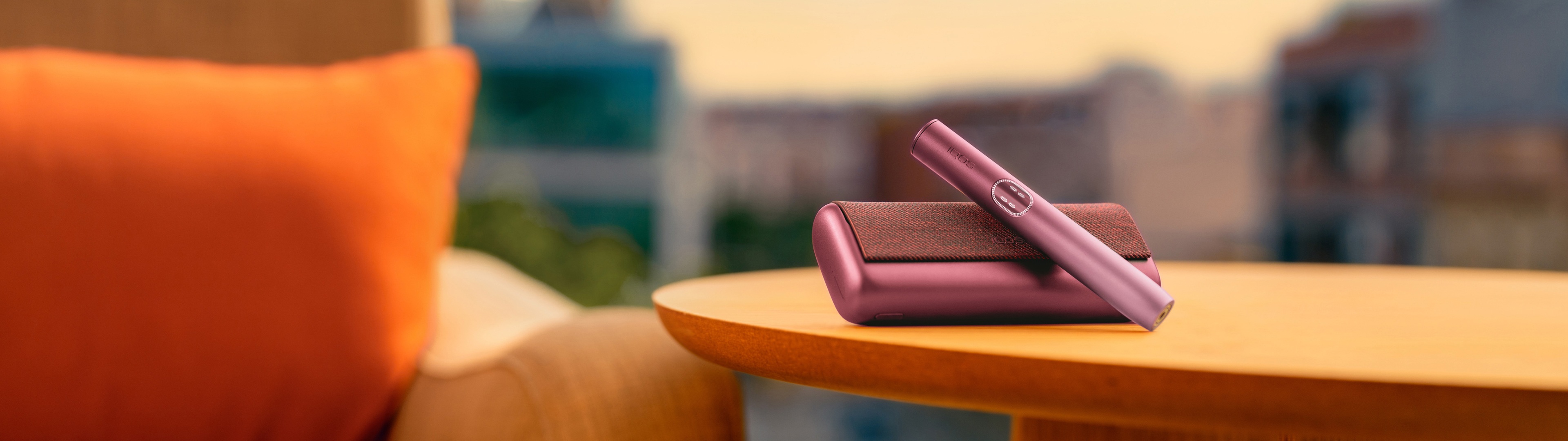 Dispositivo IQOS ILUMA i PRIME Vermelho Escuro sobre carregador de bolso, numa mesa e junto a um sofá, fundo com ambiente exterior desfocado