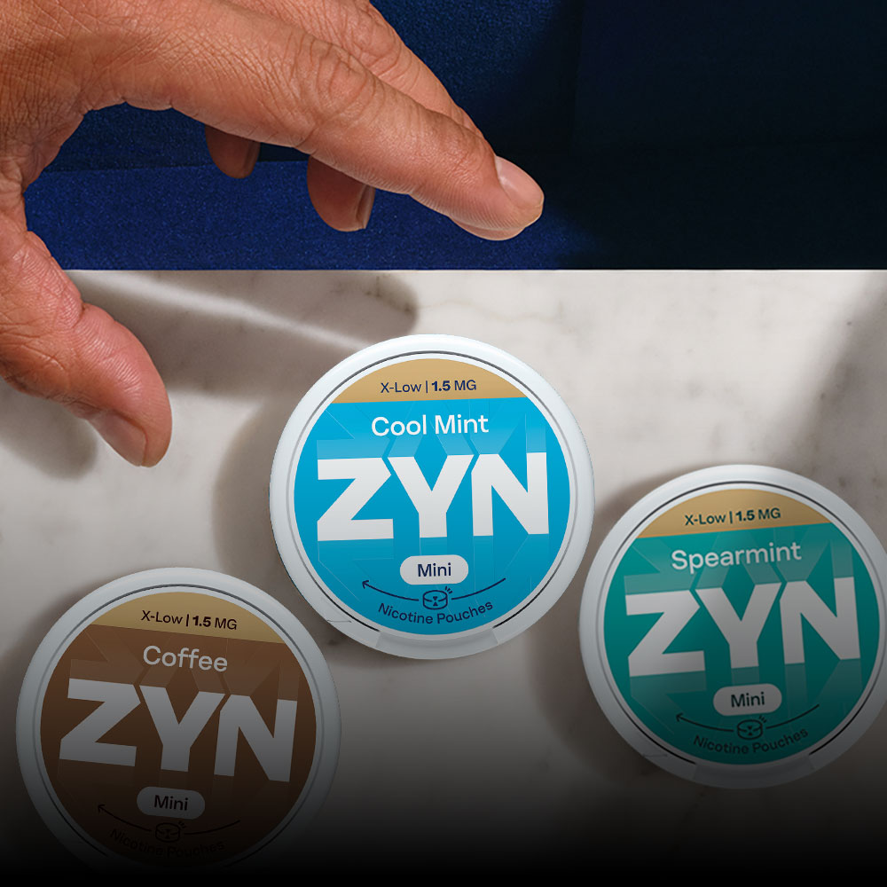 Vista de topo de uma mão de homem que se prepara para agarrar uma das três embalagens de ZYN,  ZYN Cool Mint X-Low 1,5 MG Mini,  ZYN Spearmint X-Low 1,5 MG Mini e ZYN Coffee X-Low 1,5 MG Mini, que se encontram sobre mármore branco.