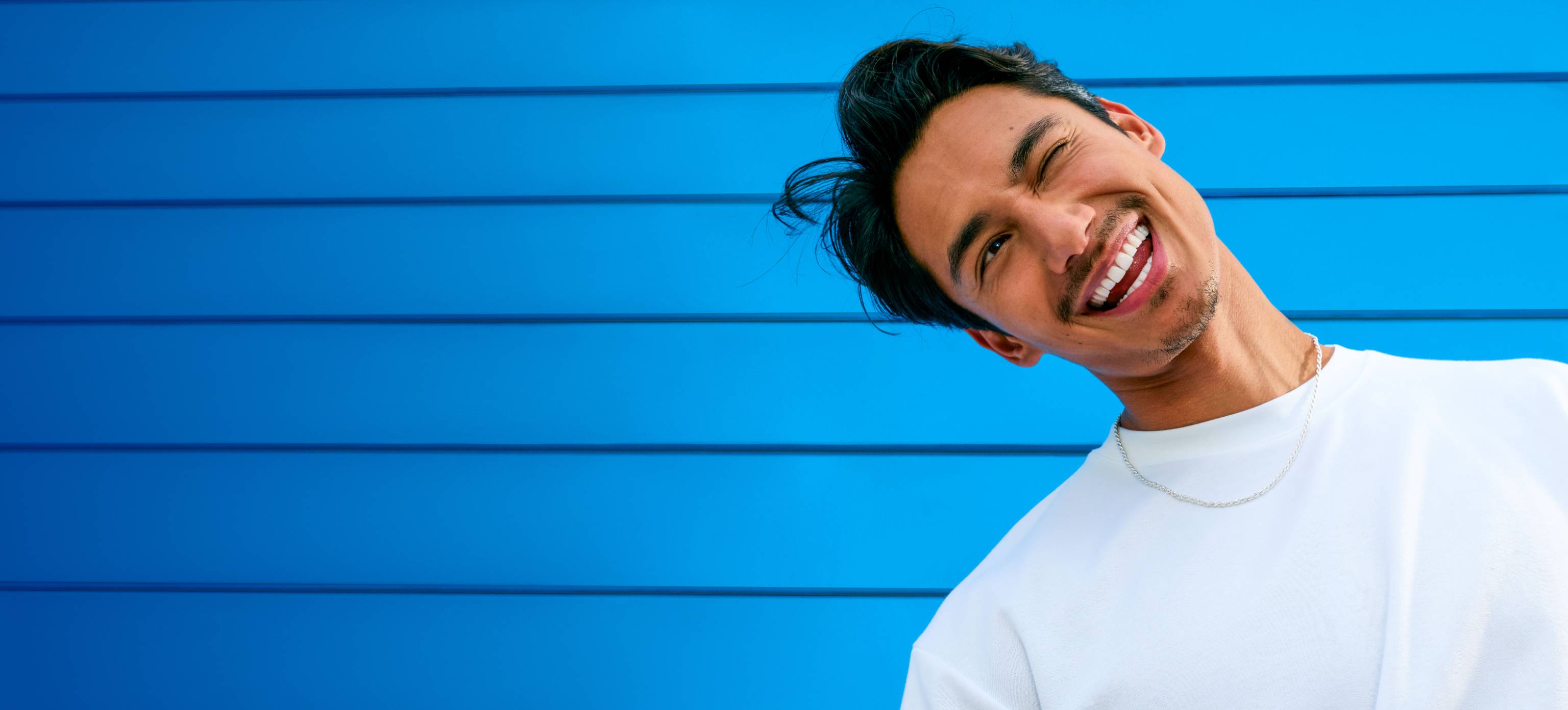 Rapaz jovem, com a cabeça inclinada para esquerda, a piscar o olho, em fundo azul.