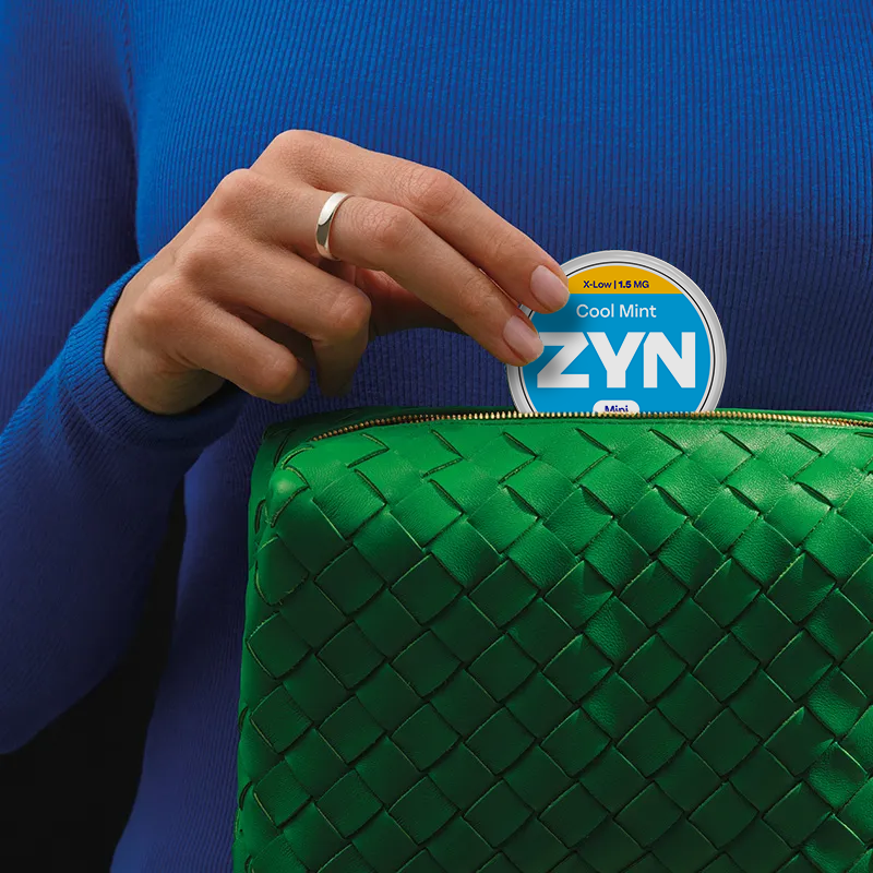 Detalhe de uma mão com aliança de mulher, de camisa azul,  a tirar uma embalagem de ZYN Cool Mint X-Low 1,5 MG Mini de uma bolsa verde.