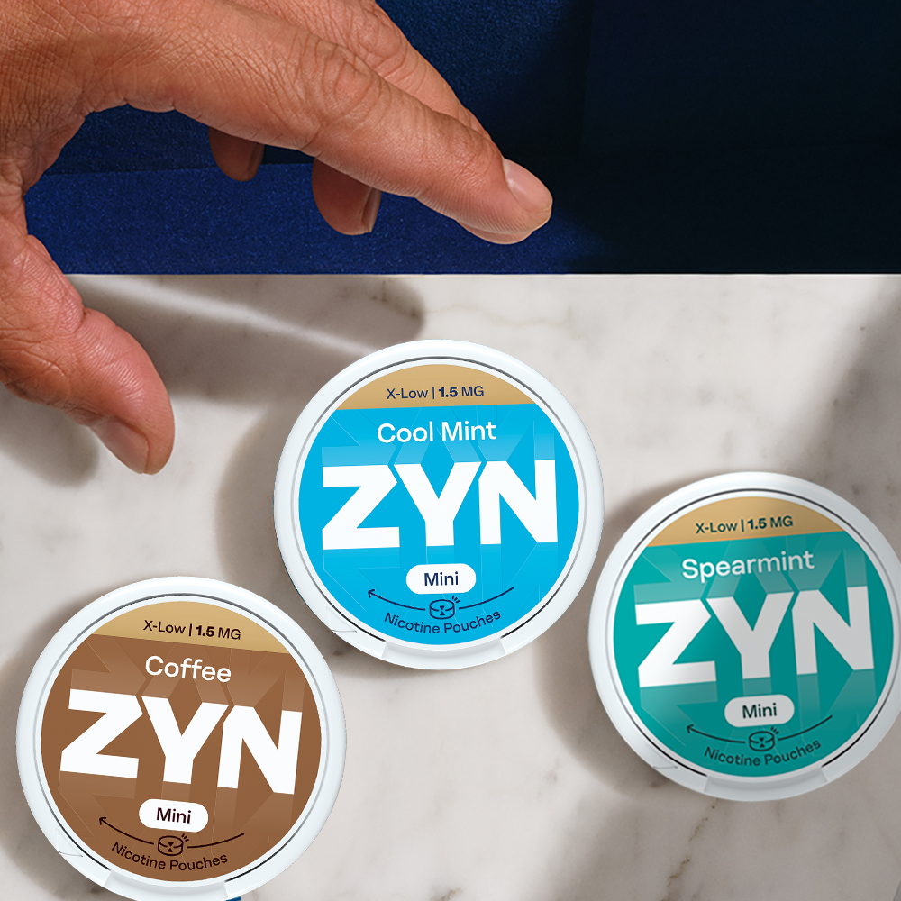 Vista de topo de uma mão de homem que se prepara para agarrar uma das três embalagens de ZYN,  ZYN Cool Mint X-Low 1,5 MG Mini,  ZYN Spearmint X-Low 1,5 MG Mini e ZYN Coffee X-Low 1,5 MG Mini, que se encontram sobre mármore branco.