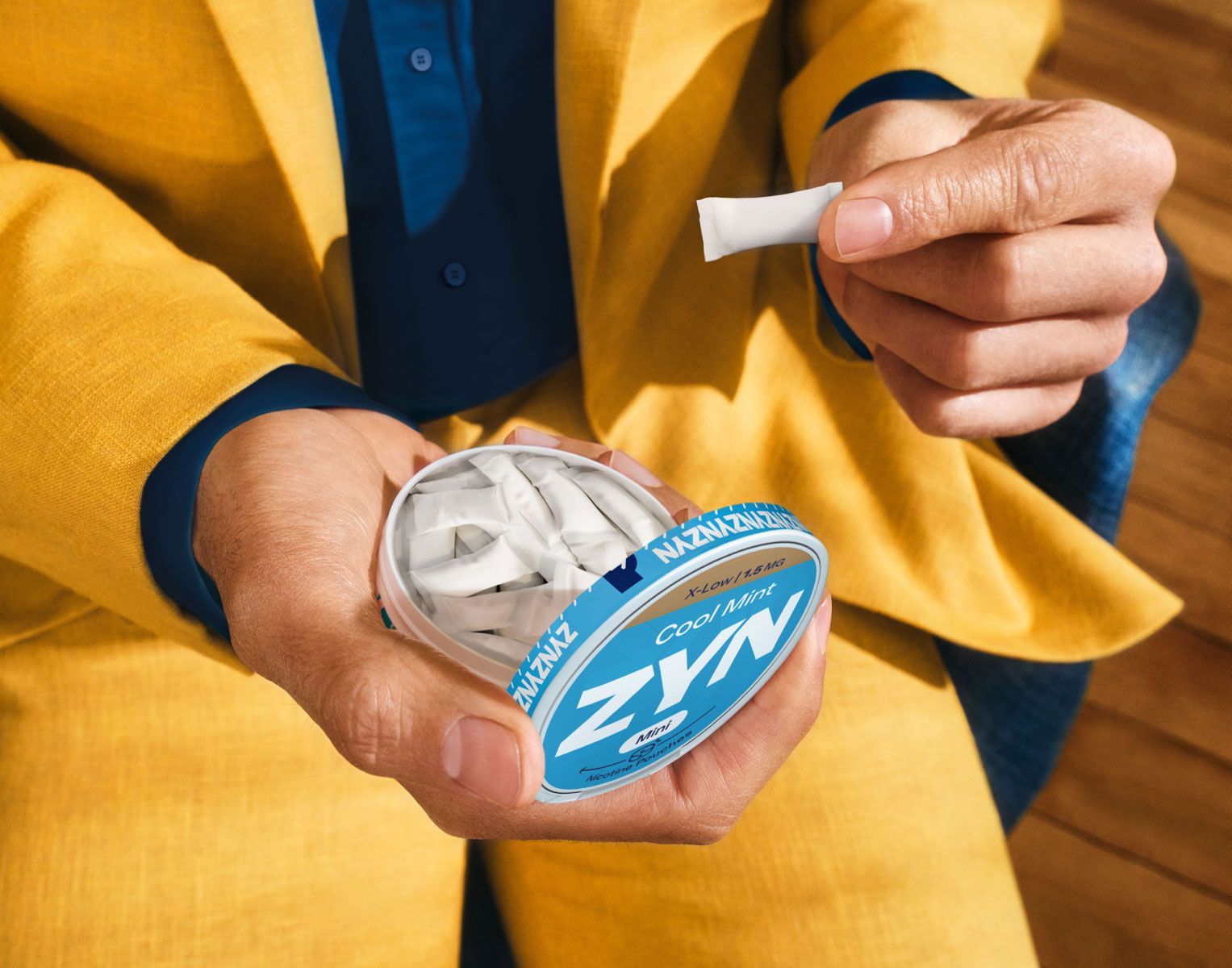 Homem de fato amarelo e camisa azul escura a segurar uma lata de ZYN aberta cheia de bolsas de nicotina numa mão e com uma bolsa de nicotina ZYN na outra mão, está a segurar esta bolsa com o dedo indicador e polegar