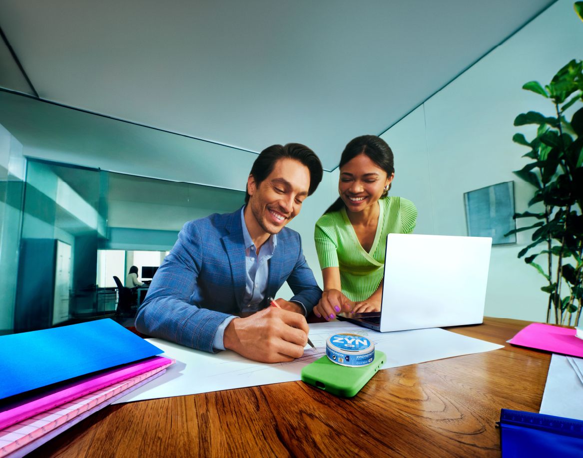Homem e mulher sentados numa secreetária em frente a um computador portátil, tiram apontamentos, com uma lata de ZYN em cima da secretária