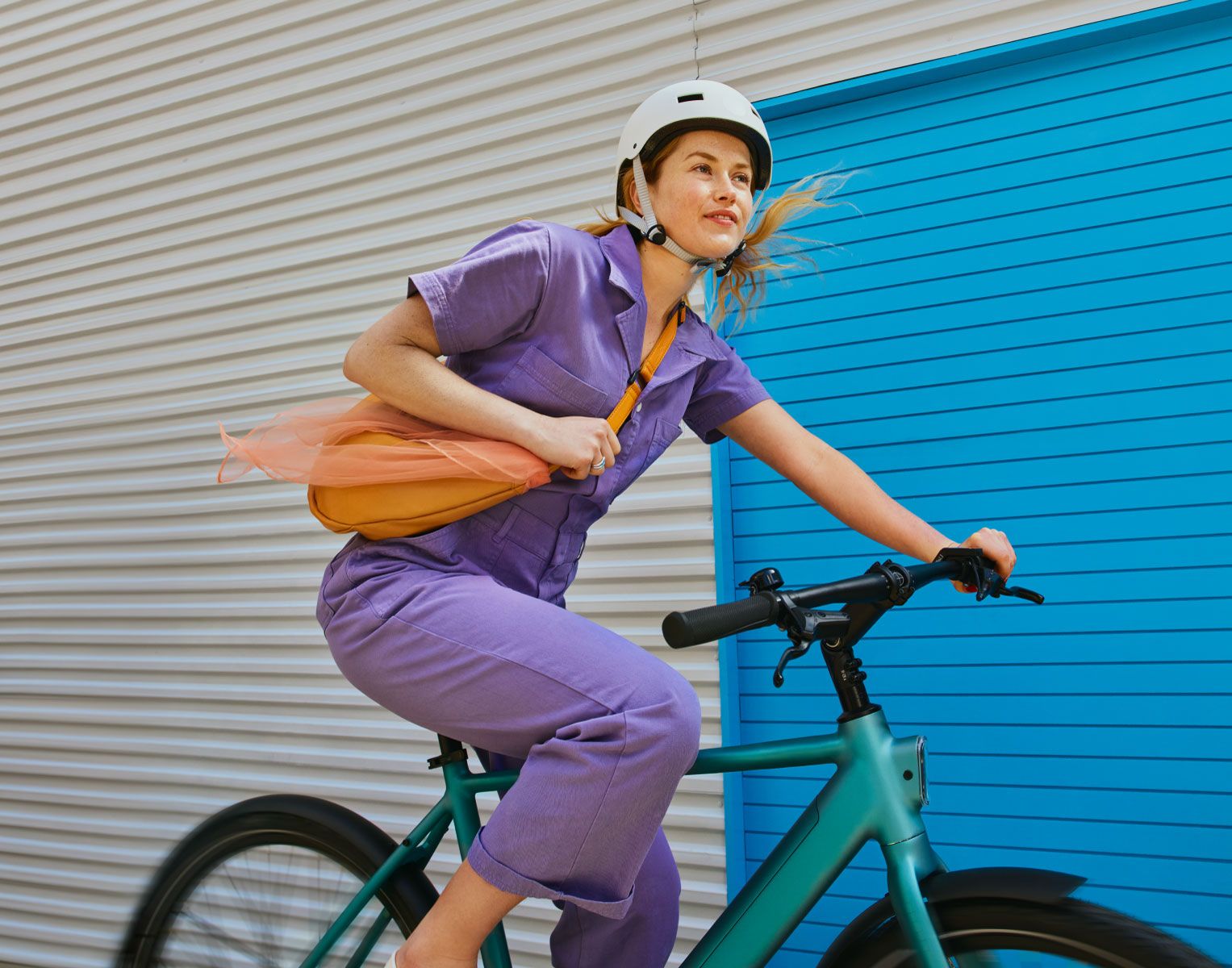 Senhora vestida com um macacão lilás a andar de bicicleta com um capacete branco