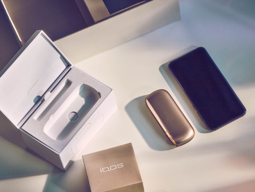 Um dispositivo IQOS Dourado fora da sua embalagem, acessórios e um telemóvel sobre uma mesa