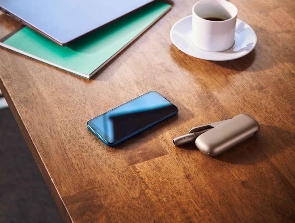 Um IQOS dourado, um telemóvel e uma chávena de café sobre uma mesa de madeira.