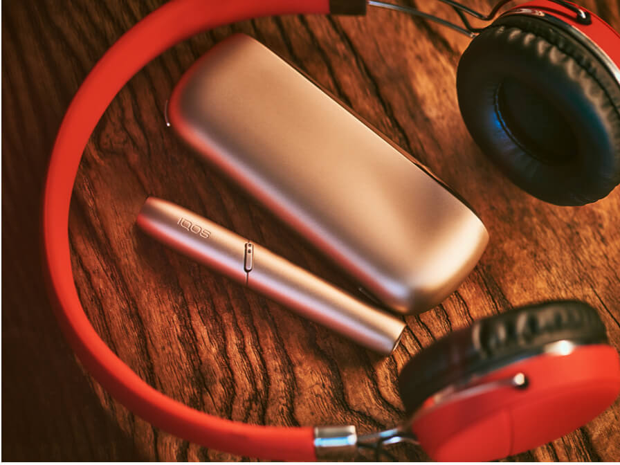 Um IQOS 3 DUO e carregador numa mesa ao lado de uns headphones vermelhos