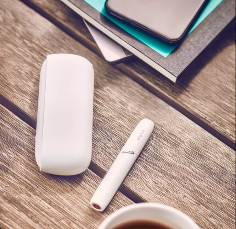 White IQOS 3 DUO charger and holder on picnic table with documents and coffee