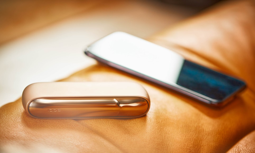 Um IQOS 3 DUO dourado e um telemóvel num sofá.