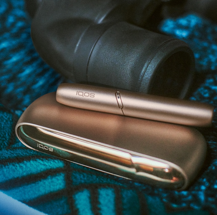 Brilliant gold IQOS 3 DUO with HEETS stick and charger next to binoculars on a blue blanket